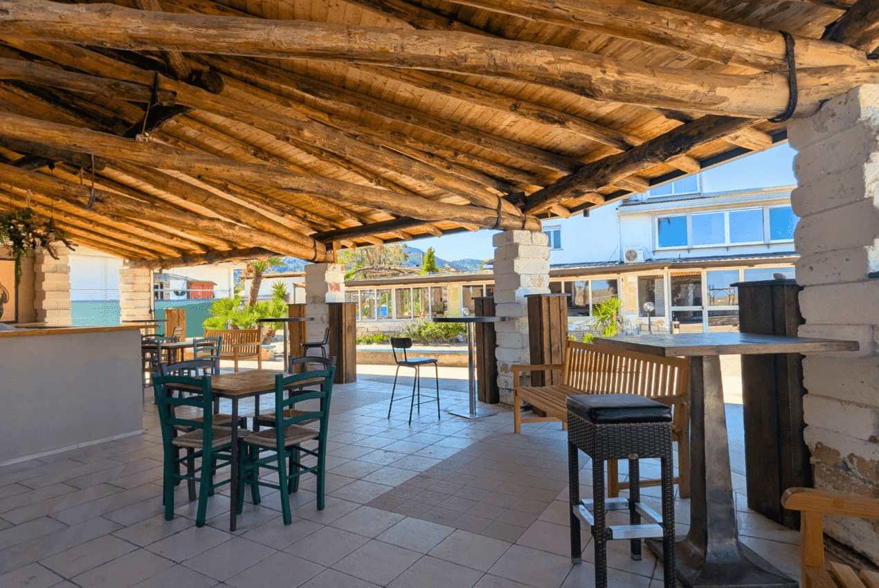 Area bar coperta del Patti Beach Resort con soffitto in travi di legno rustico, tavoli in legno e sedie colorate ideale per aperitivi a Patti