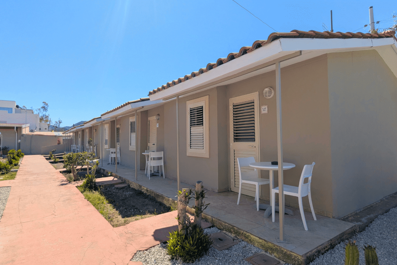 Infila di bungalow con tetto in tegole rosse e patio privato con tavolo e sedie, circondati da un giardino verde in un resort a Patti, Sicilia