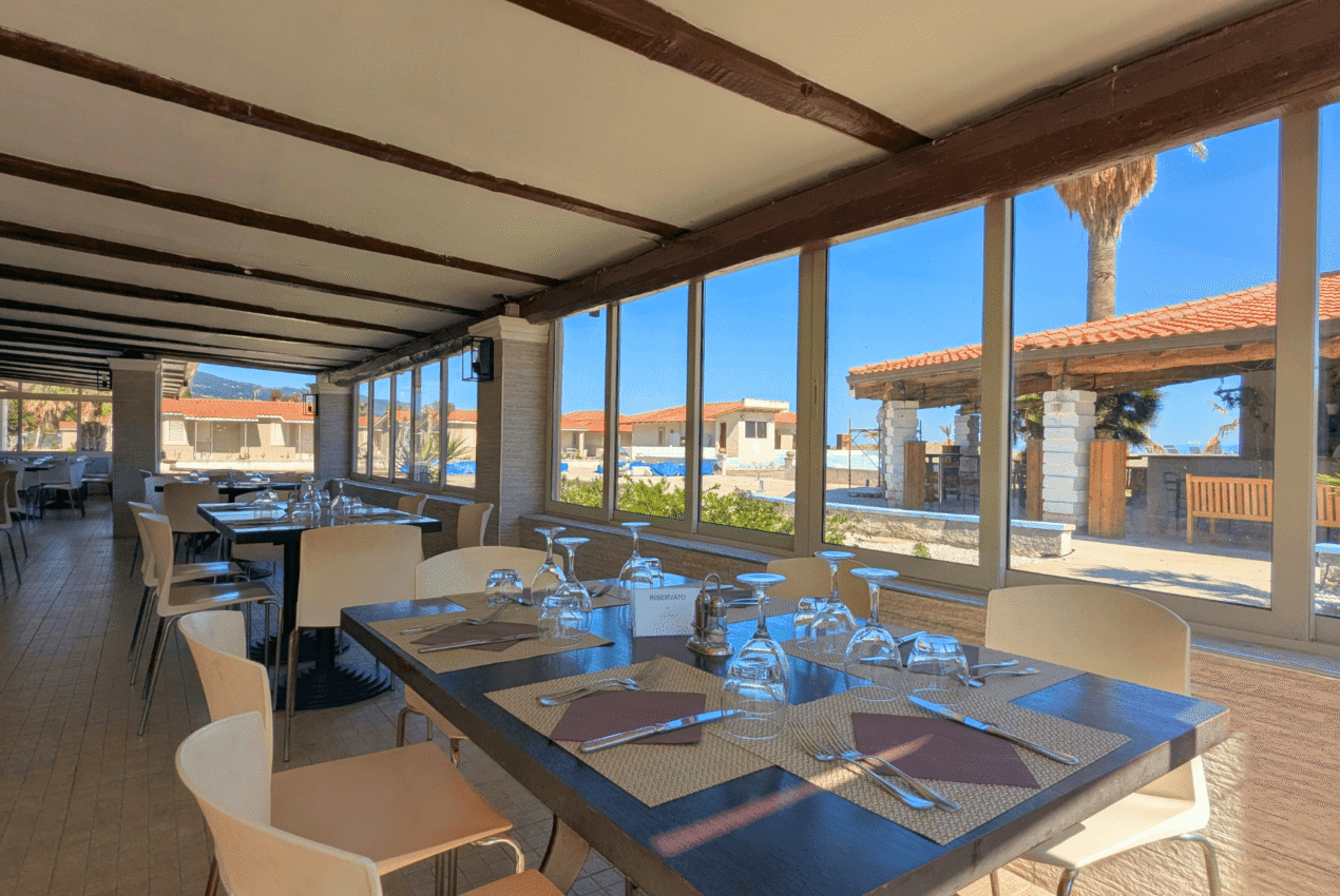 Tavolo riservato con calici e mise en place moderna presso il ristorante del Patti Beach Resort, con vista sugli spazi esterni del villaggio