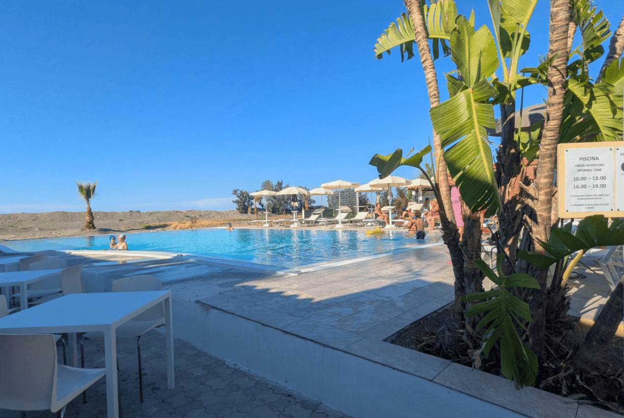 Area relax con tavolini bianchi e piante tropicali a bordo piscina del Patti Beach Resort