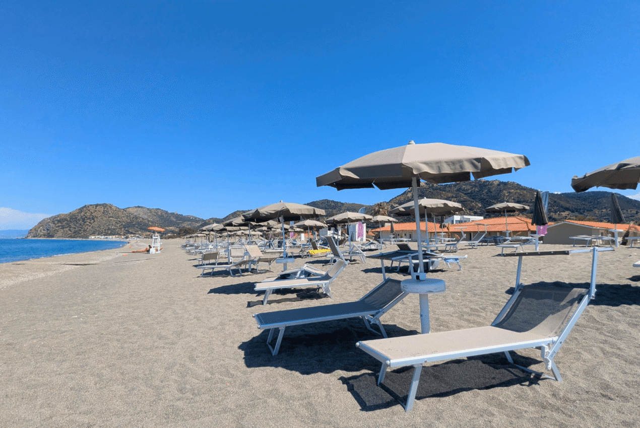 Lettini prendisole e ombrelloni beige sulla spiaggia del Patti Beach Resort con vista sul promontorio circostante